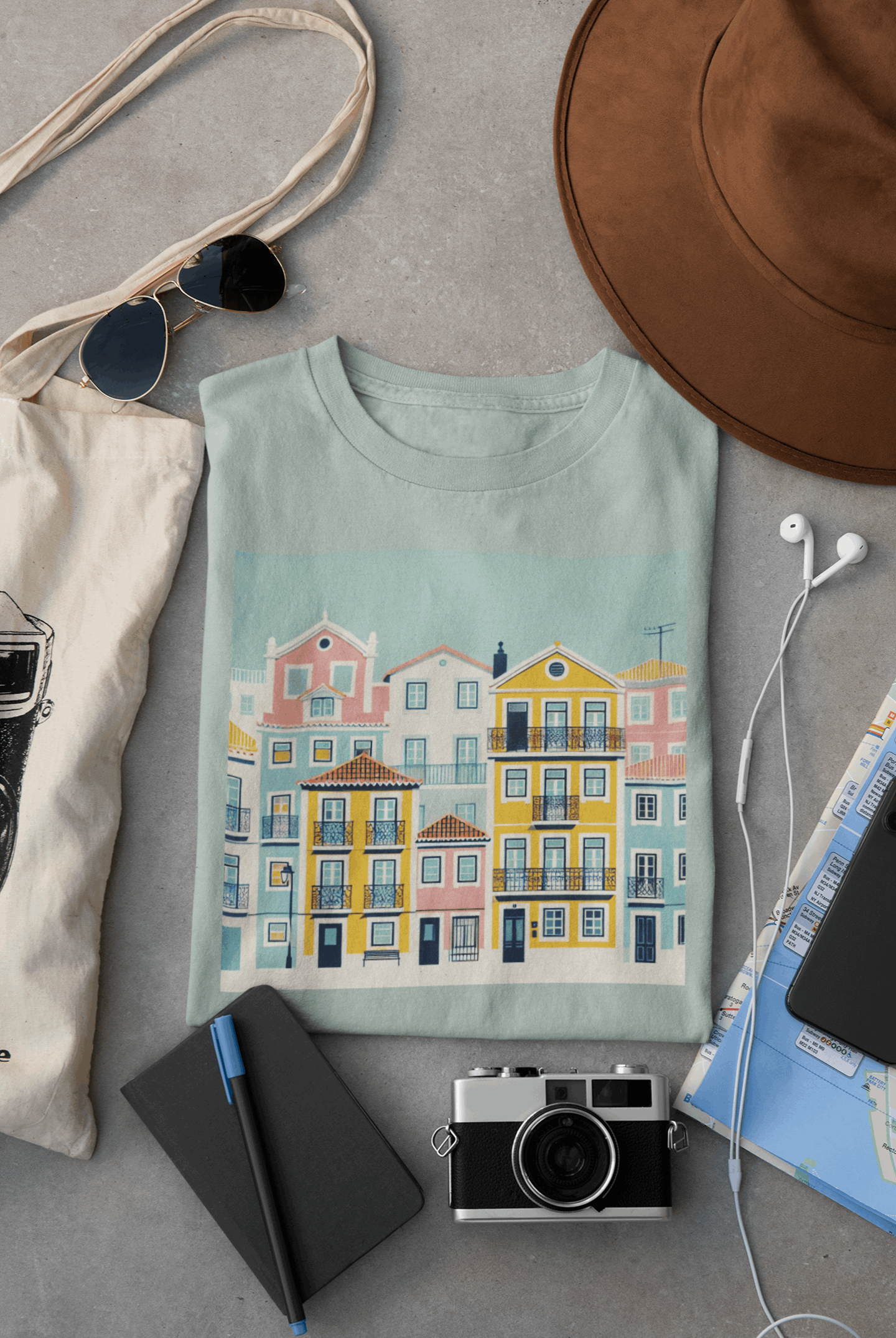 Folded light blue unisex T-shirt with Lisbon Alfama houses design, surrounded by travel essentials. Organic cotton and eco-friendly fashion.