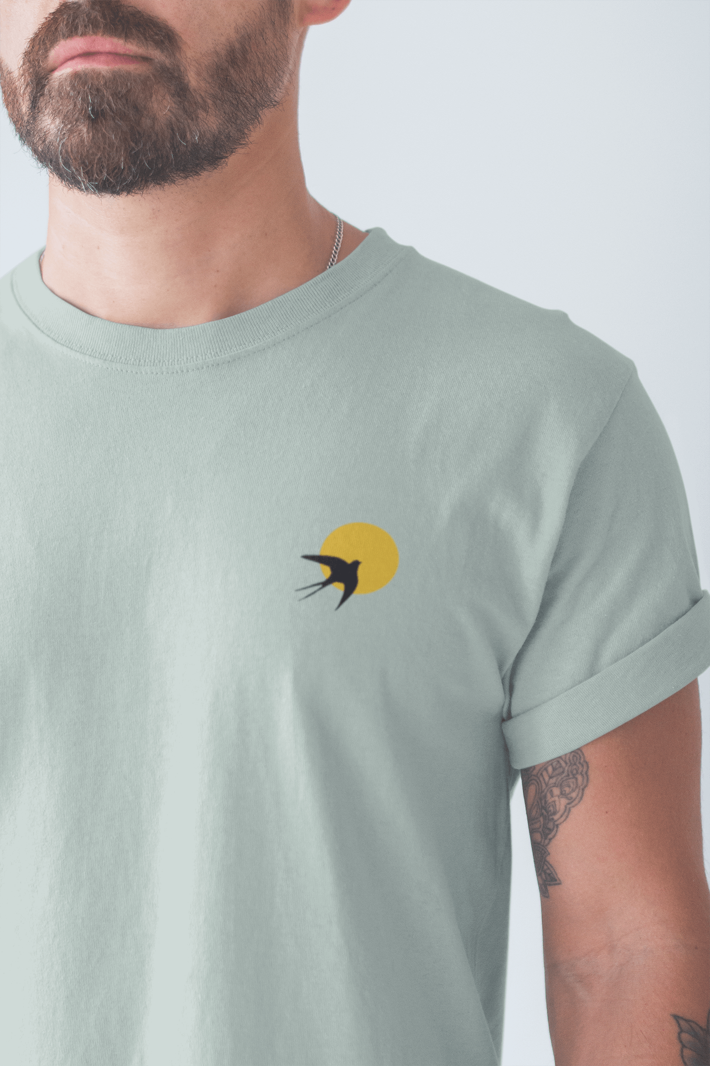 Close-up of a sage green Andorinha T-Shirt with a swallow and sun logo. A unisex organic cotton tee inspired by Portugal's heritage.