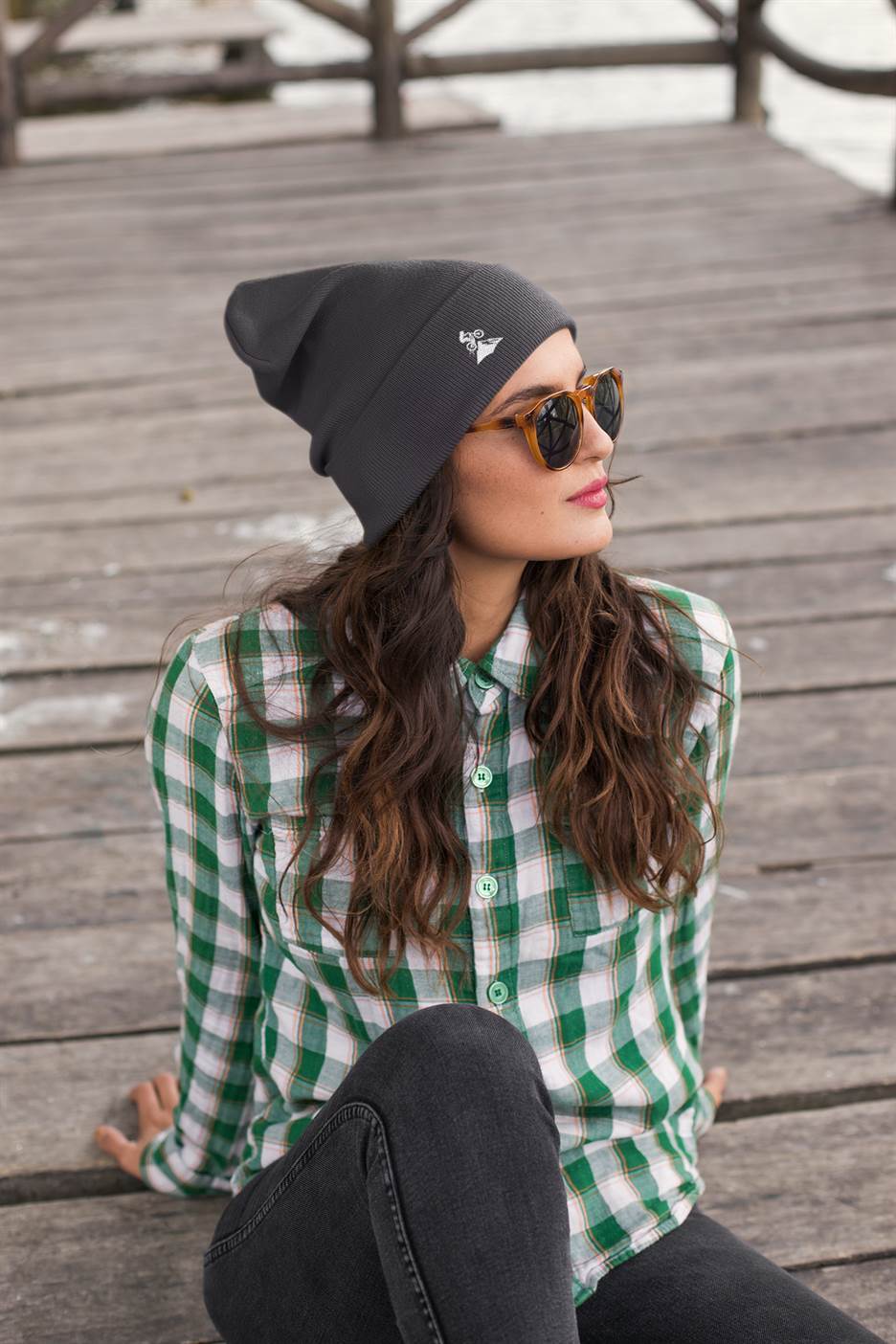 Dark gray Madeira Beanie with embroidered mountain bike logo, worn by a woman in sunglasses on a wooden dock. Stylish and warm for adventures.