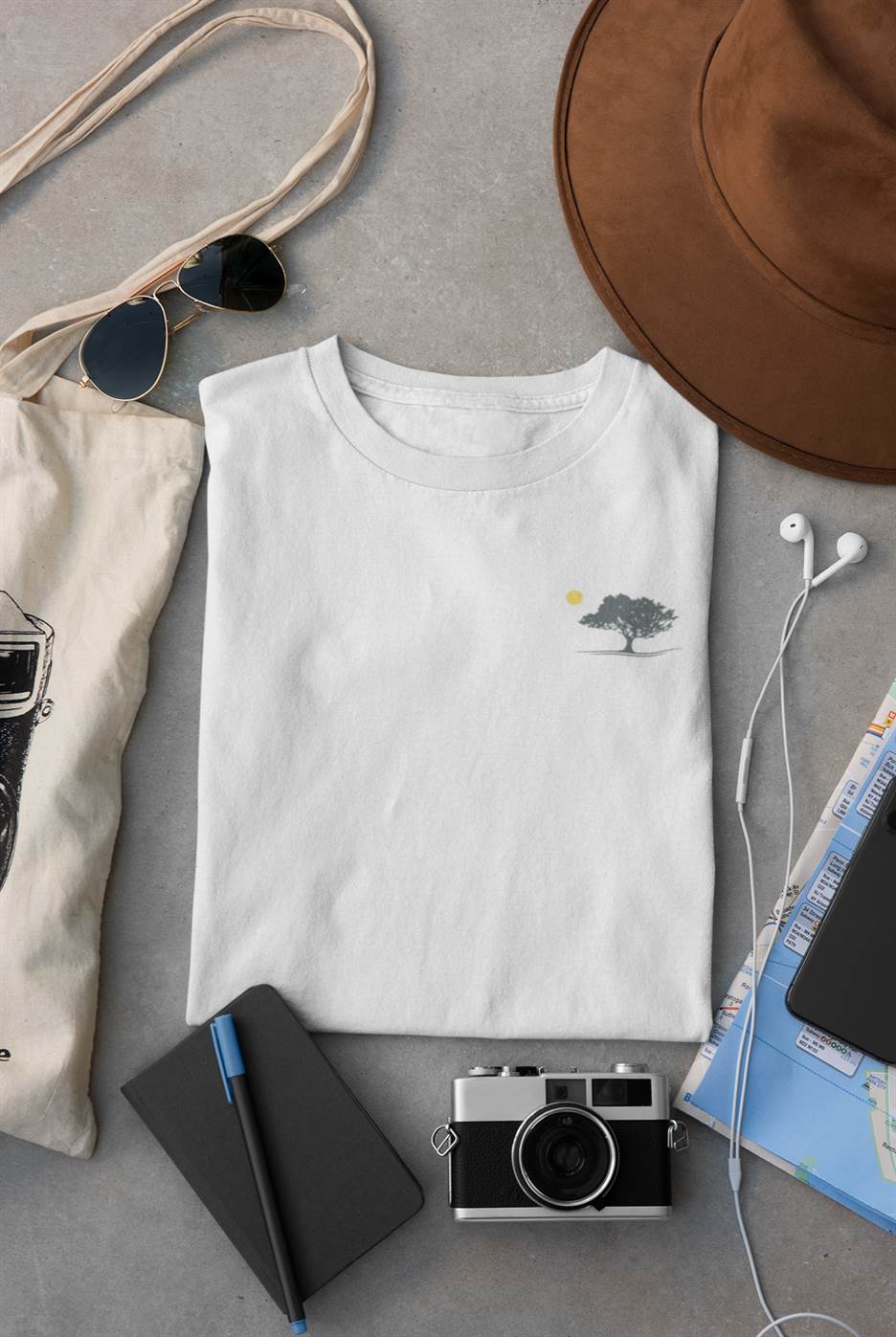 Folded white organic T-shirt with a minimalist oak tree and sunset design, styled with travel essentials. Perfect for sustainable and casual wear.