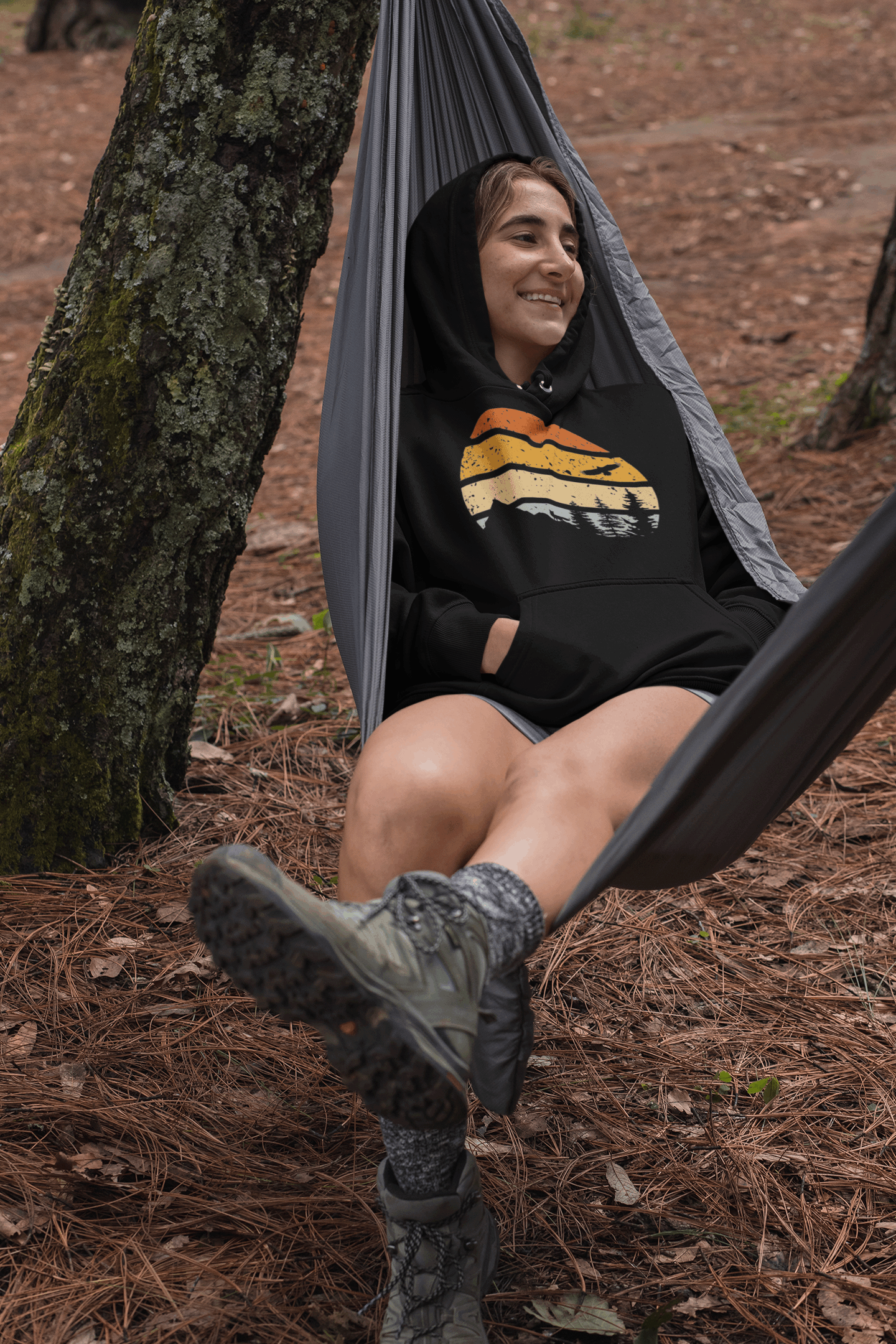 Black organic hoodie with a retro sunset and mountain design, worn by a woman relaxing in a hammock in a forest setting.
