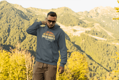 Men’s organic adventure hoodie in blue with a retro sunset print, styled for outdoor exploration in a mountainous landscape.