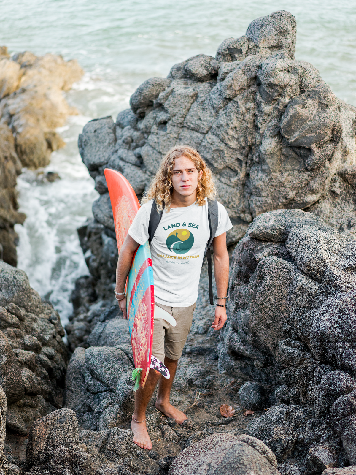 Woman wearing organic cotton white T-shirt with retro surf graphic and "Land & Sea" text, holding a pink surfboard on rocky.