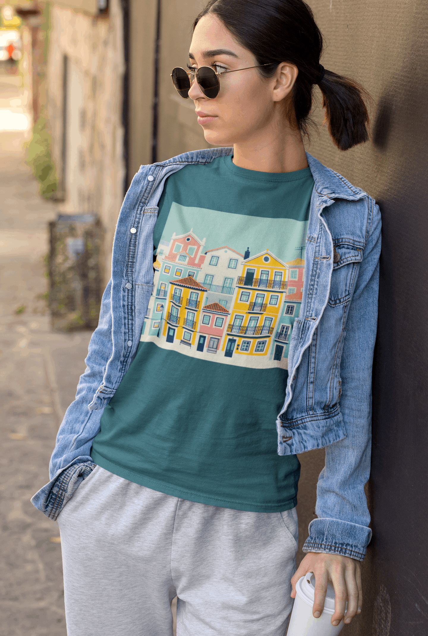 Woman in a teal unisex T-shirt with Lisbon Alfama houses design, styled with a denim jacket. Eco-friendly and trendy organic cotton apparel.
