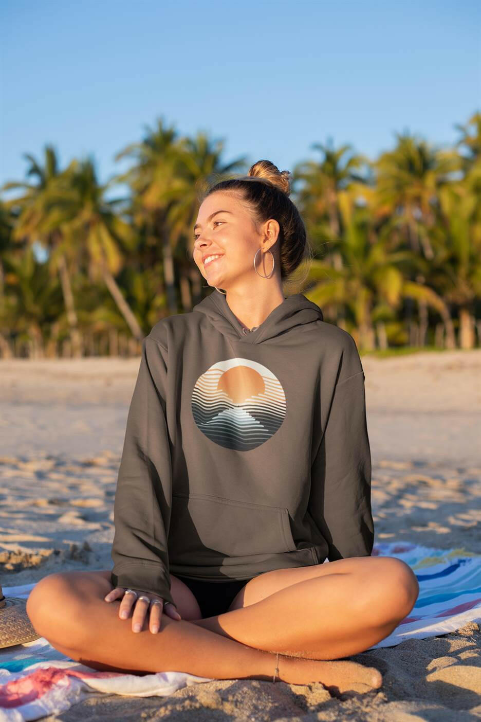 Dark green Nazaré vintage hoodie with a sunset print, worn by a woman sitting on the beach. Cozy and eco-friendly surfwear for any occasion.