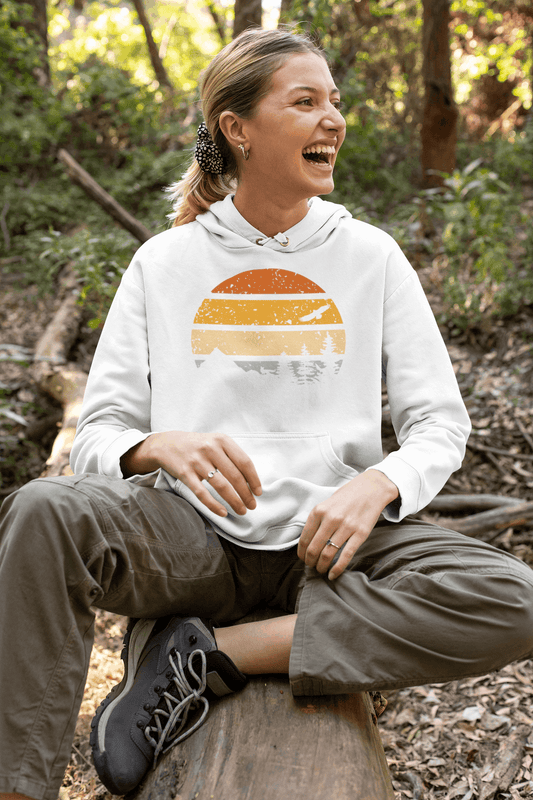 Unisex organic cotton hoodie with a vintage sunset and nature design, worn by a woman sitting on a log in a forest setting.