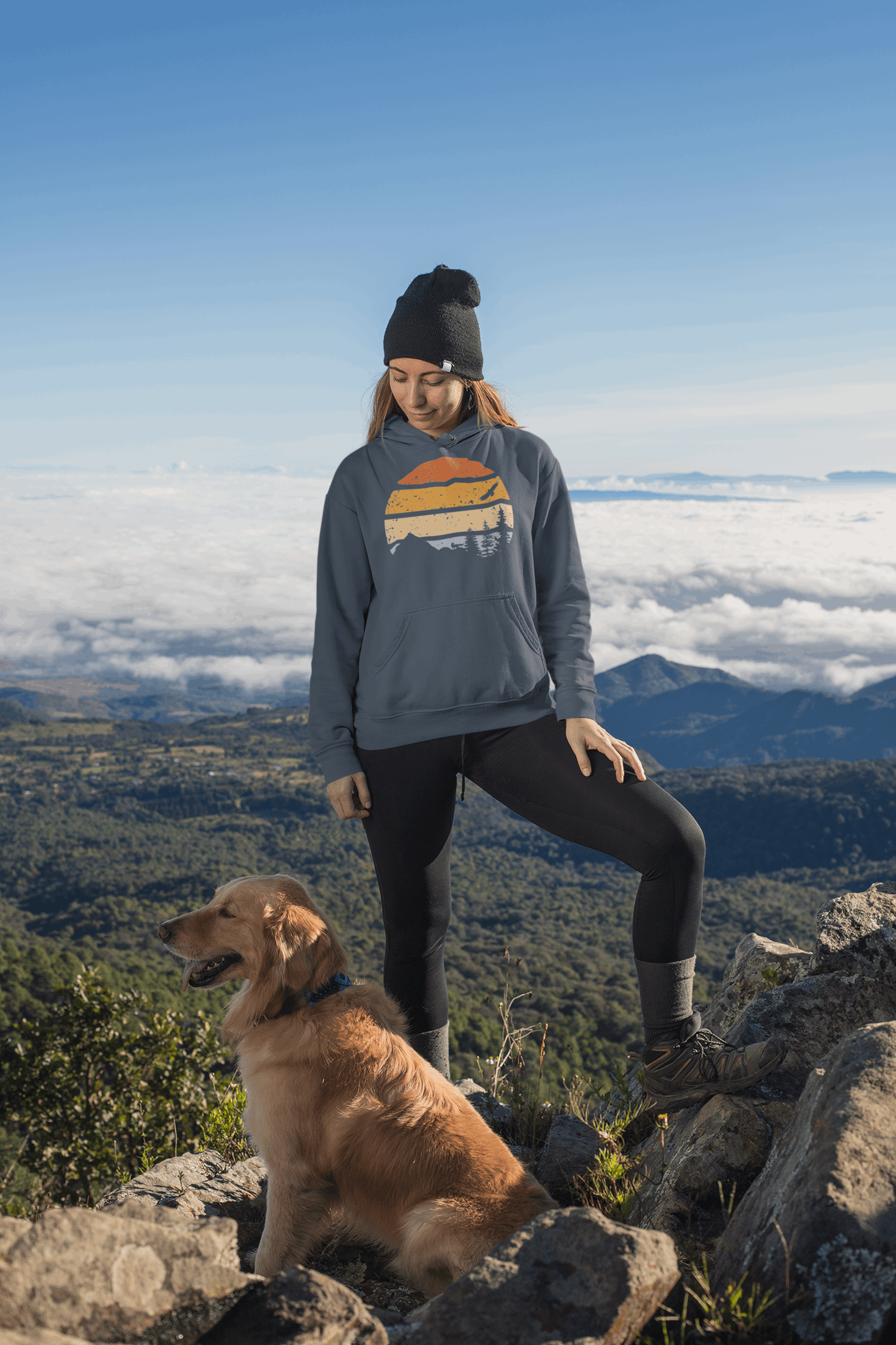 Unisex adventure hoodie in blue with a retro nature print, worn by a woman hiking with her dog above the clouds.
