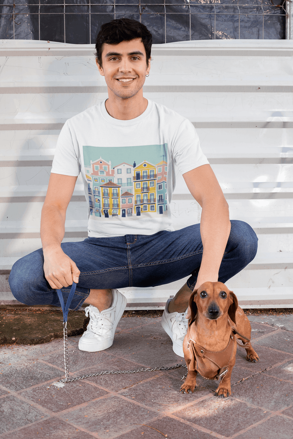 Smiling man in a white unisex T-shirt with Lisbon Alfama houses design, crouching with a dachshund. Organic cotton and eco-friendly style.