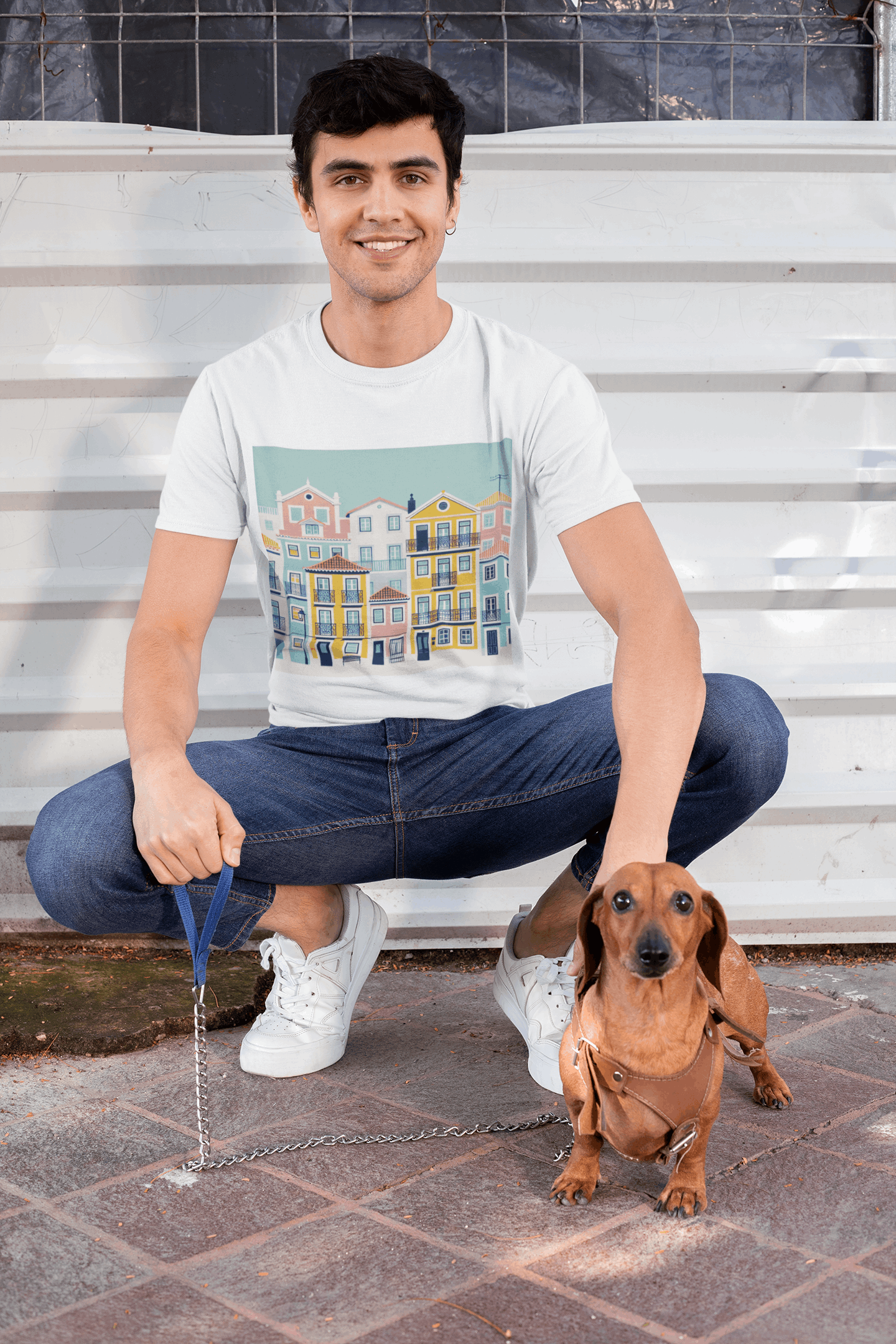 Smiling man in a white unisex T-shirt with Lisbon Alfama houses design, crouching with a dachshund. Organic cotton and eco-friendly style.