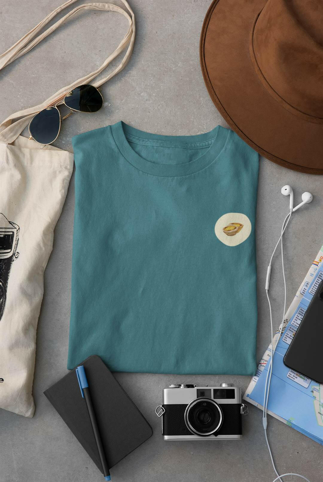 Pastel de Nata T-Shirt in teal, folded with travel essentials. Organic cotton, minimalist Lisbon style for men.