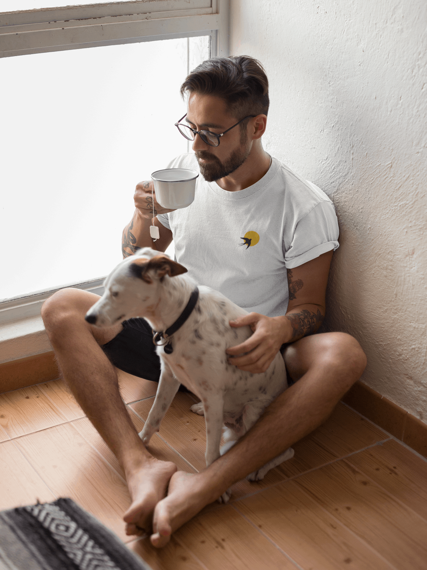 Man wearing a white Andorinha T-Shirt with a swallow logo, sitting with a dog while drinking tea. A minimalist unisex organic cotton tee.