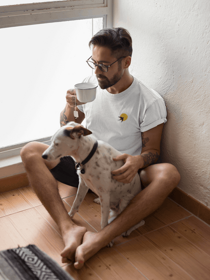 Man wearing a white Andorinha T-Shirt with a swallow logo, sitting with a dog while drinking tea. A minimalist unisex organic cotton tee.