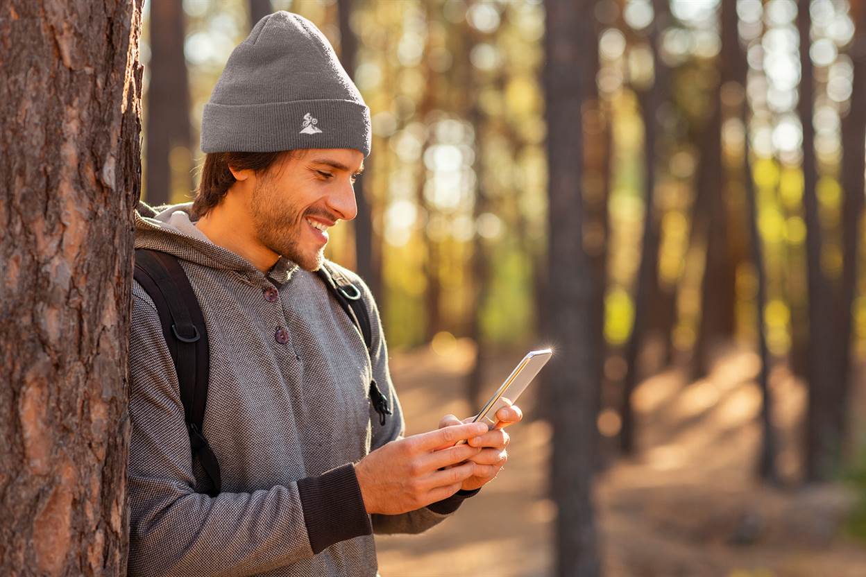 Gray Madeira Beanie with embroidered mountain bike logo, worn by a man using his phone in a forest. A great choice for adventurers.