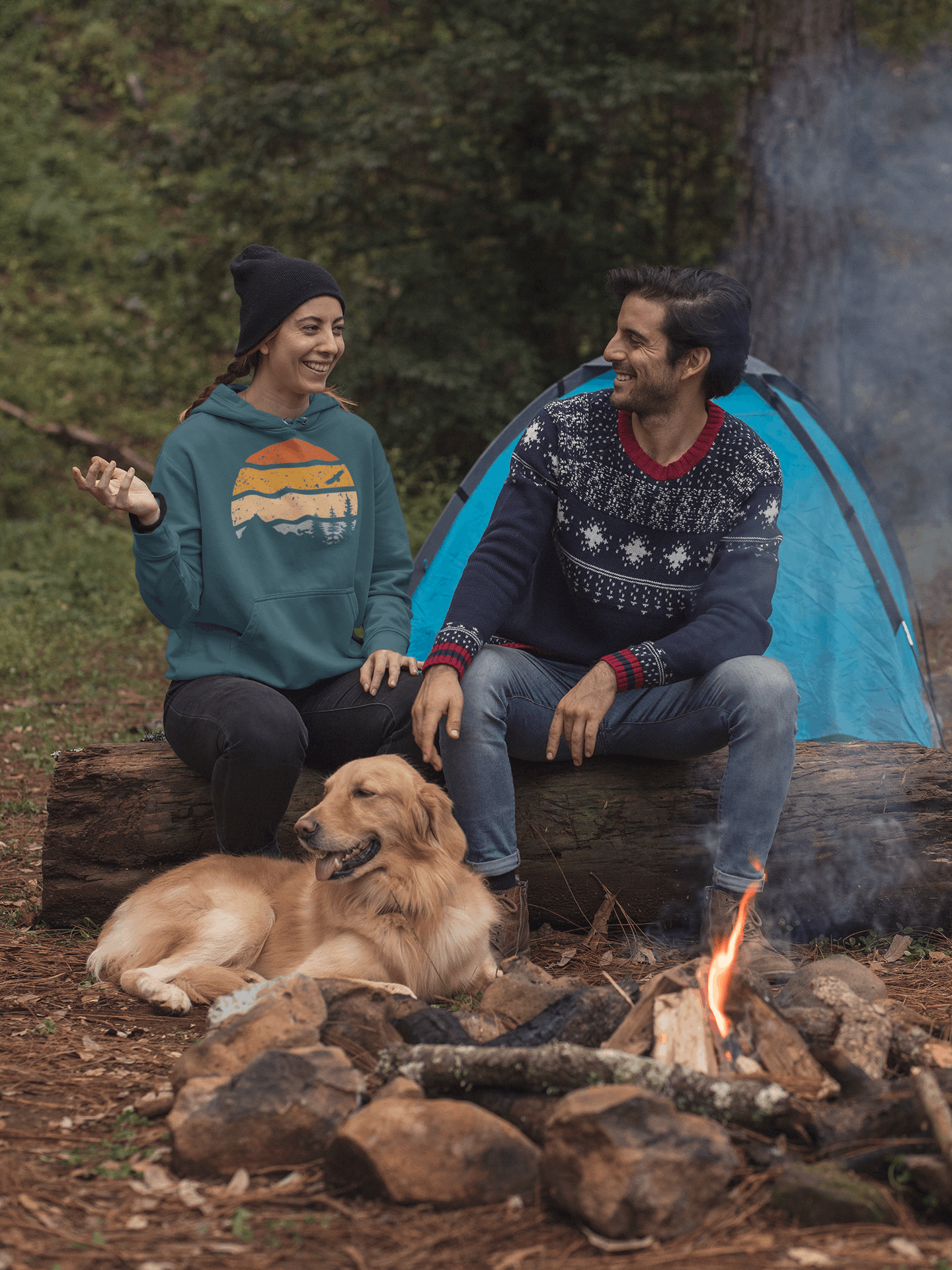 Teal organic cotton hoodie with a retro sunset print, worn by a woman sitting by a campfire with a dog and a companion.