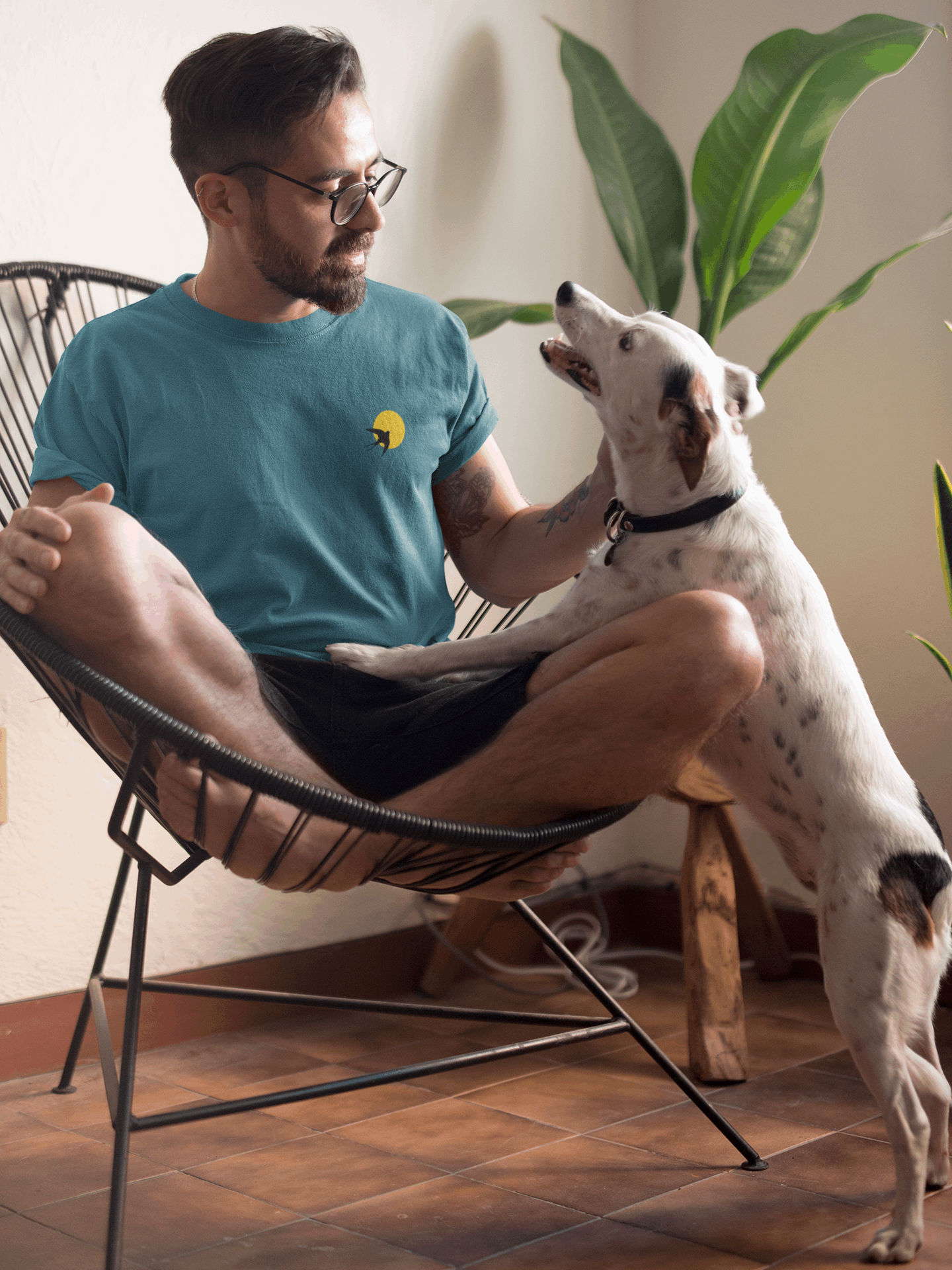 Man in a blue Andorinha T-Shirt sitting in a chair, interacting with a dog. A unisex organic cotton tee with a minimalist swallow and sun logo.