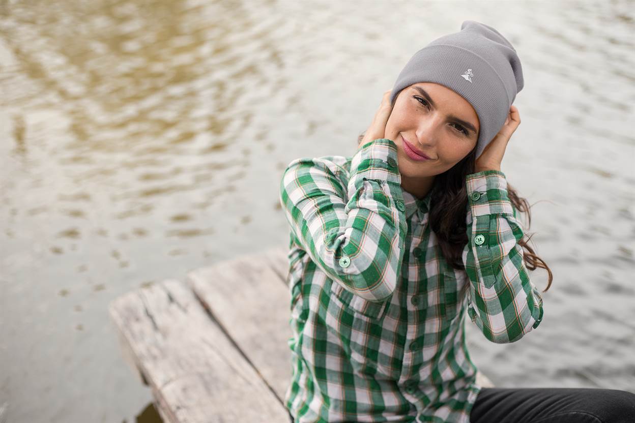Light gray Madeira Beanie with embroidered mountain bike logo, worn by a woman sitting by the water. Ideal for outdoor and adventure lovers.