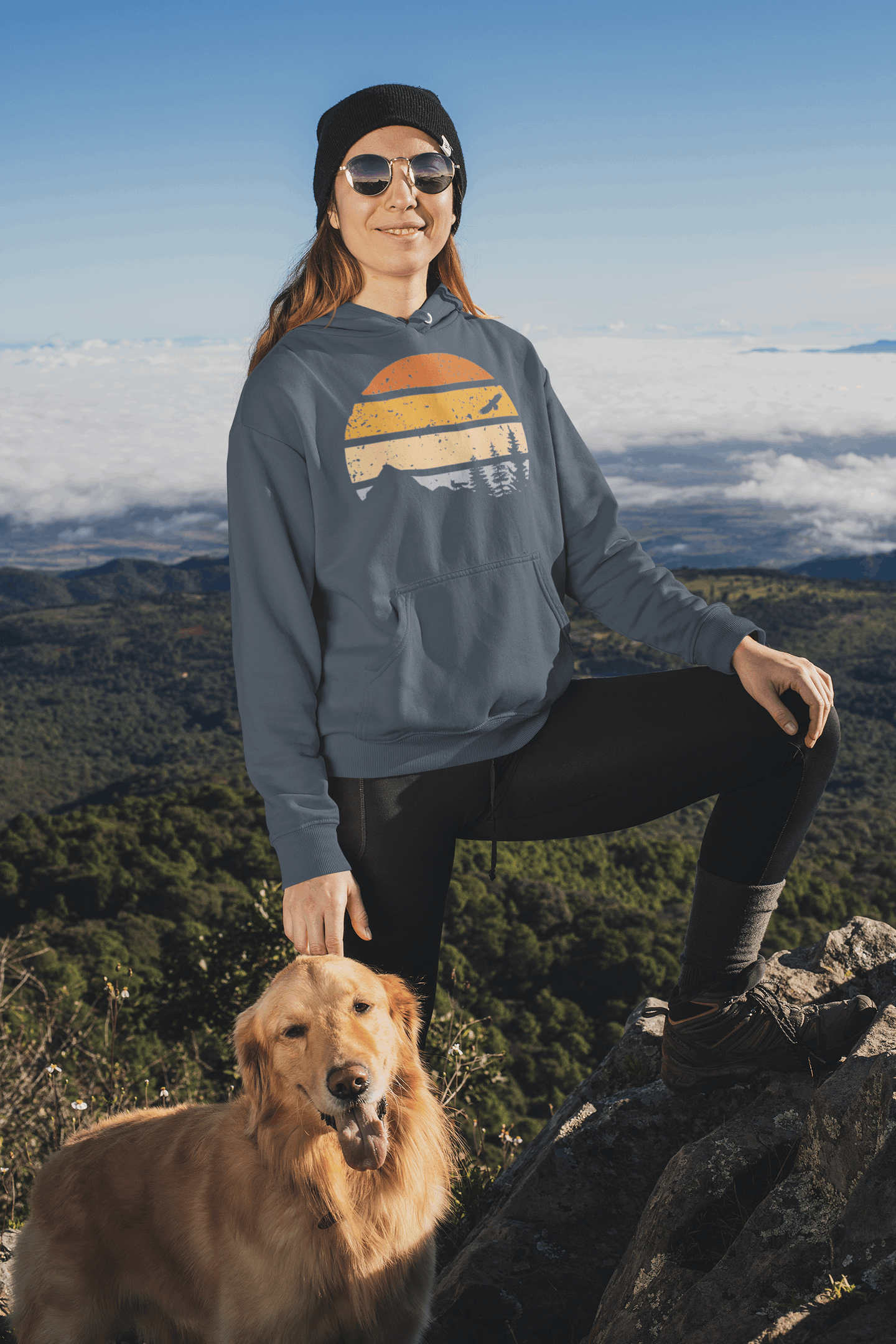 Blue organic cotton hoodie with a vintage sunset and wildlife print, worn by a woman hiking with her dog on a mountain trail.