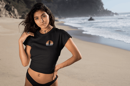 Woman in a black cropped Nazare Surf Minimal Organic T-shirt with a vintage surf logo, standing on a sandy beach. A stylish and sustainable surf-inspired tee.