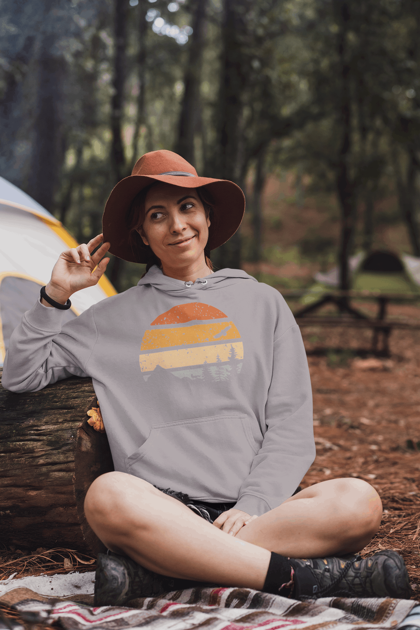 Light gray organic hoodie with a vintage sunset and forest design, worn by a woman relaxing at a campsite.