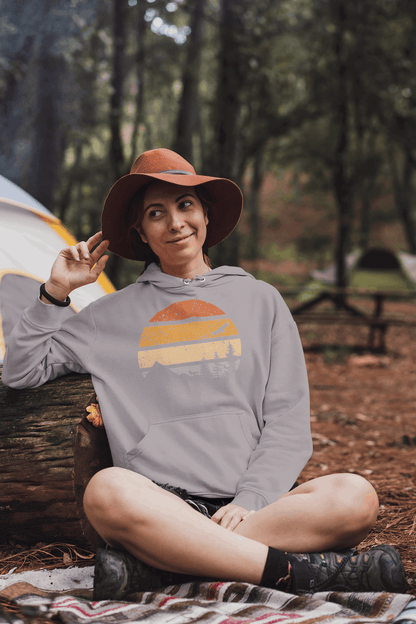 Light gray organic hoodie with a vintage sunset and forest design, worn by a woman relaxing at a campsite.