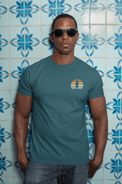Man wearing a teal Nazare Surf Minimal Organic T-shirt with a vintage surf logo, posing against blue patterned tiles. A classic and eco-friendly unisex tee.