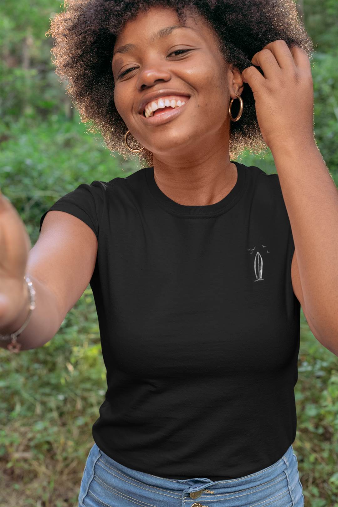 Black Ericeira Surf Organic T-Shirt for women with a minimalist surfboard design, styled casually with jeans for a natural and joyful outdoor look.
