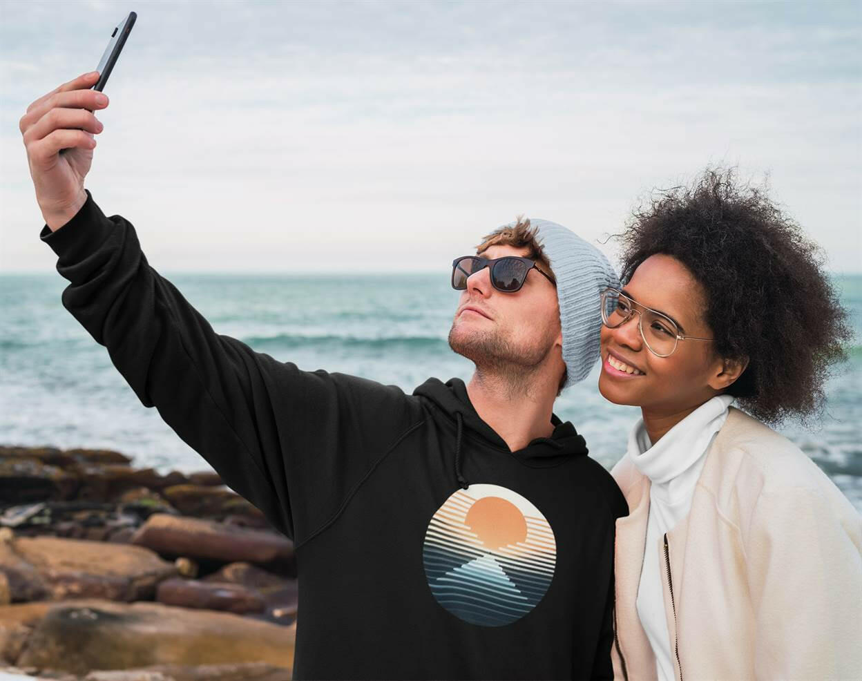 Black Nazaré vintage hoodie with a sunset surf print, worn by a couple taking a selfie by the ocean. Stylish and eco-friendly unisex hoodie.