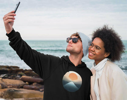 Black Nazaré vintage hoodie with a sunset surf print, worn by a couple taking a selfie by the ocean. Stylish and eco-friendly unisex hoodie.