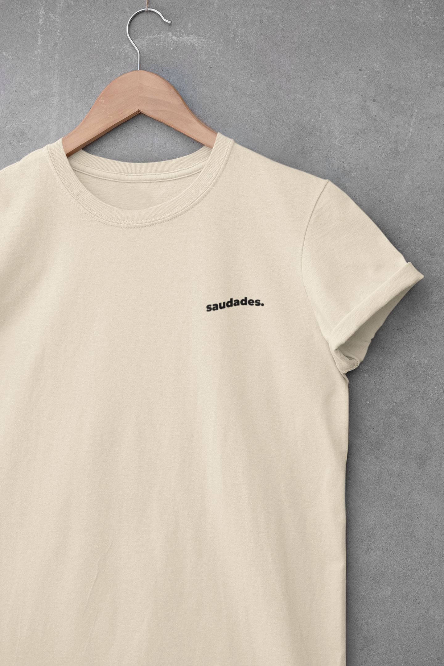 Beige t-shirt with 'saudades.' text on a hanger against a gray background