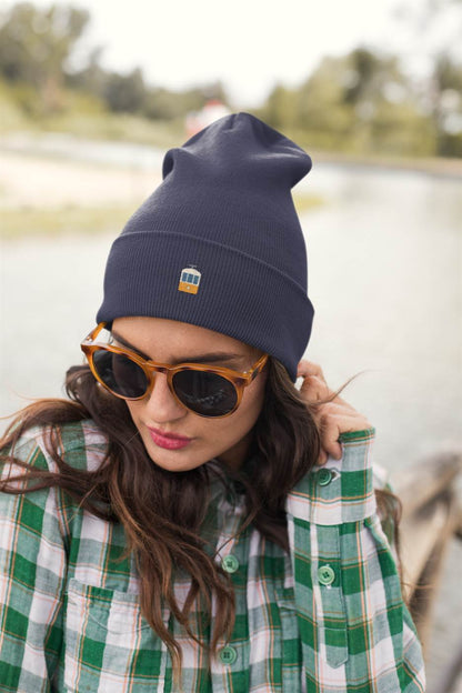 Navy blue Lisbon Electrico Beanie with embroidered tram detail, styled with sunglasses and a plaid shirt. A warm and stylish turbo acrylic hat.